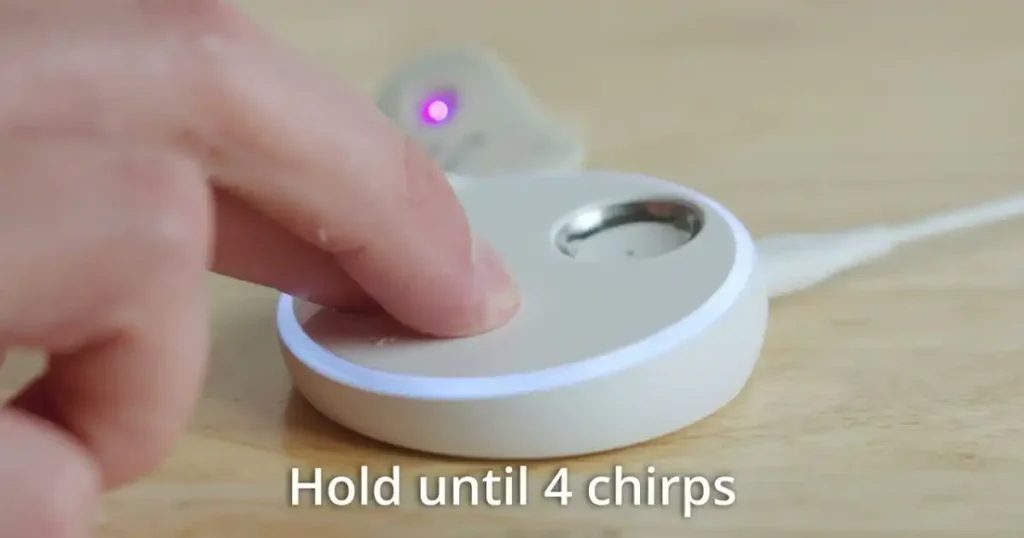 A person's finger pressing a button on the Owlet Sock base station. The text "Hold until 4 chirps" is displayed on the image.